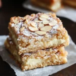 A close-up of almond croissant cookie bars topped with sliced almonds and powdered sugar on a parchment-lined tray.